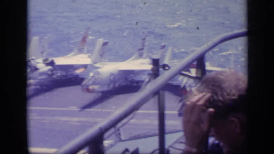 A man at the right edge looks toward white jet fighters lined on a carrier flight deck, with sea visible beyond them.