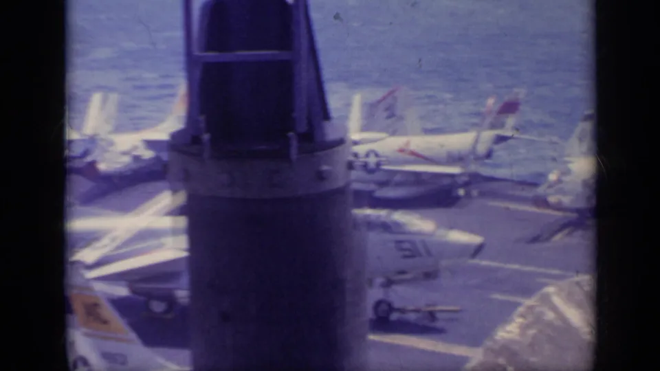 A dark deck structure divides the frame as parked jet aircraft and flight-deck markings remain visible in the background.