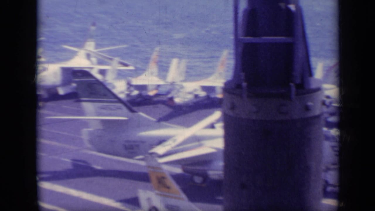 Distant view of carrier deck with parked military aircraft and dark window or frame bars in foreground.