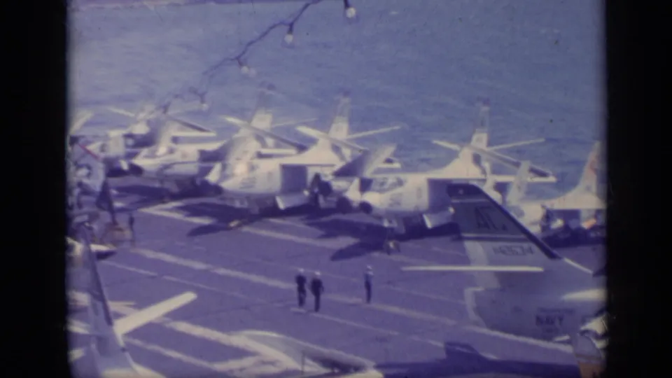 1962 Italy-area naval scene with multiple military aircraft parked on a carrier deck and crew figures visible between them.
