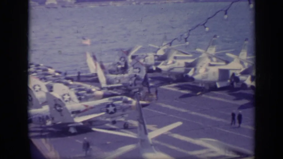 Oblique 1962 8mm view of a naval aircraft carrier deck with parked planes, visible markings, and crew figures near the deck edge.