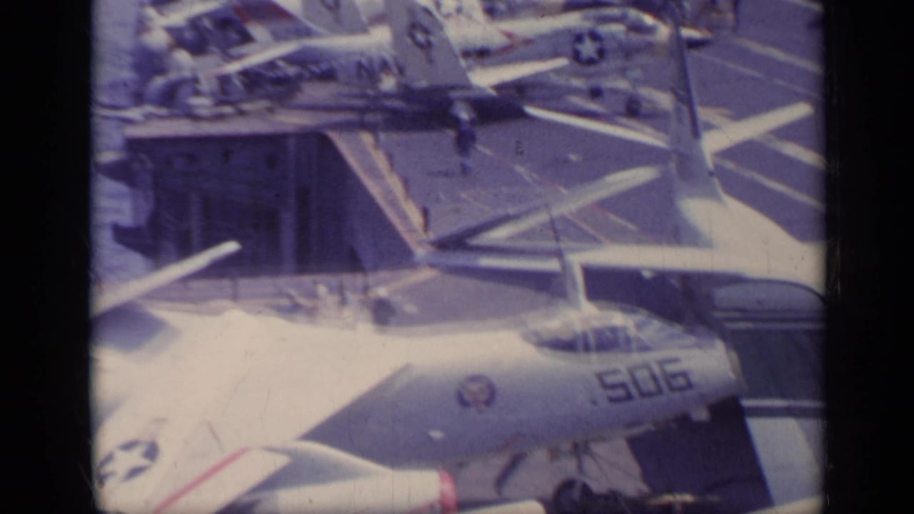 Parked U.S. Navy jet aircraft on a carrier deck with deck markings and small crew figures.