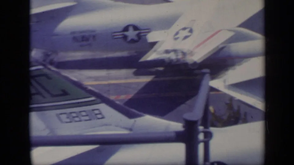 1962 Italy-associated 8mm frame showing the side of a U.S. Navy jet on a carrier deck, with star insignia and railing crossing the foreground.