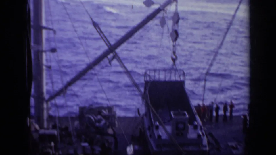 A smaller boat appears to be hoisted by crane lines beside a naval vessel at sea in 1962 Italy footage.