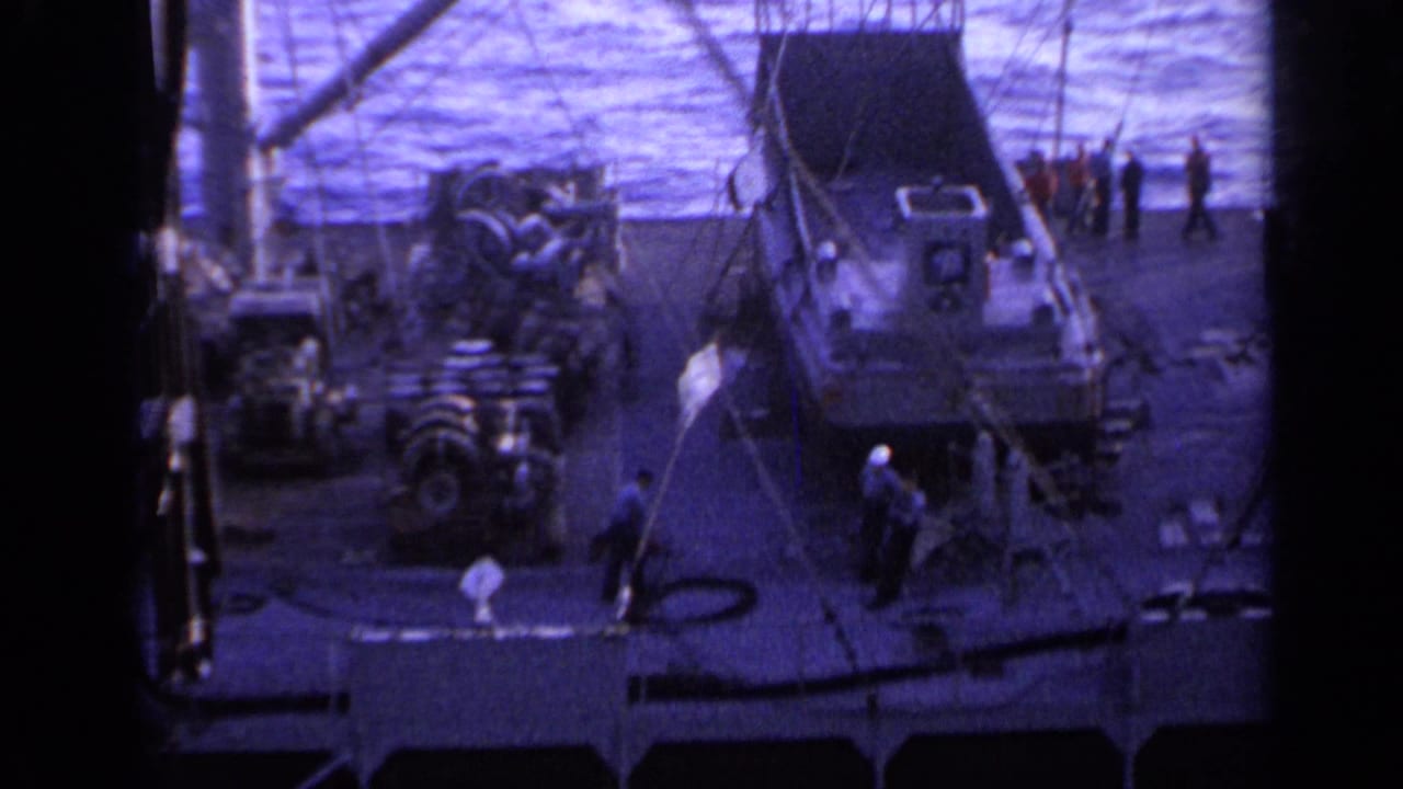 1962 Italy 8mm frame showing a ship deck near open water, with crew members, crane lines, and a small boat or tender on deck.