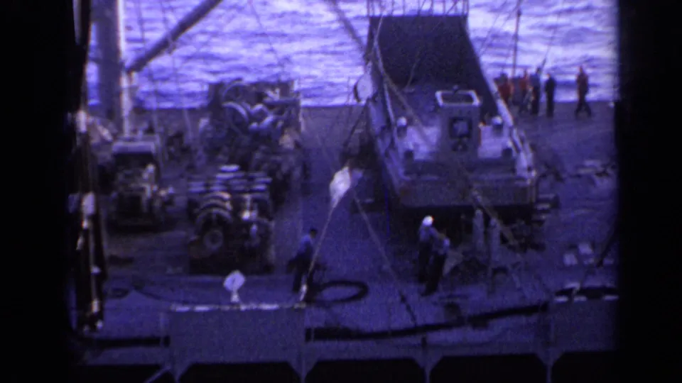 1962 Italy 8mm frame showing a ship deck near open water, with crew members, crane lines, and a small boat or tender on deck.