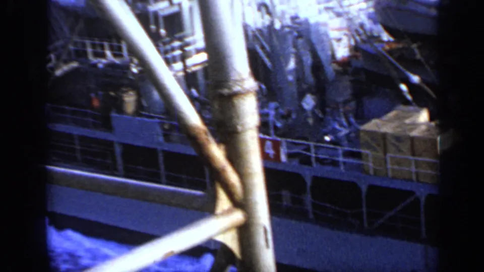 1962 Italy 8mm frame with cargo boxes, ship railings, crew-like figures in the distance, and large crane supports in the foreground.