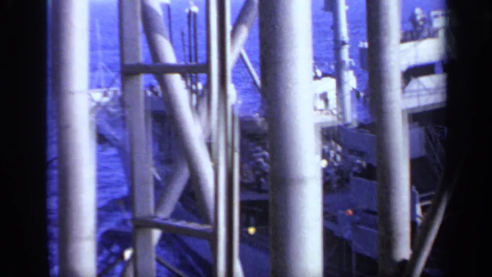 A white vessel at sea is viewed through vertical ship supports, with blue water and deck details visible in the background.