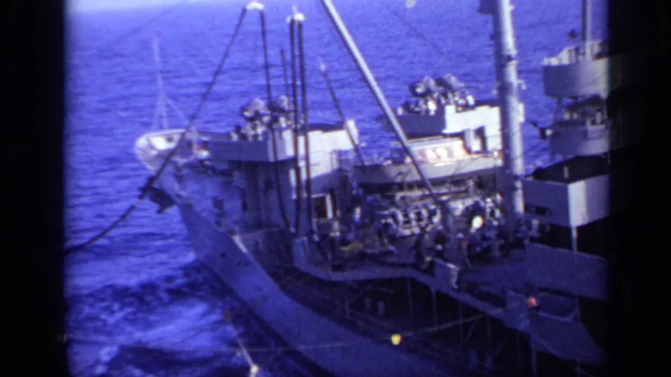 A white ship with visible deck machinery and small crew figures is filmed at sea from another vessel in 1962 near Italy.