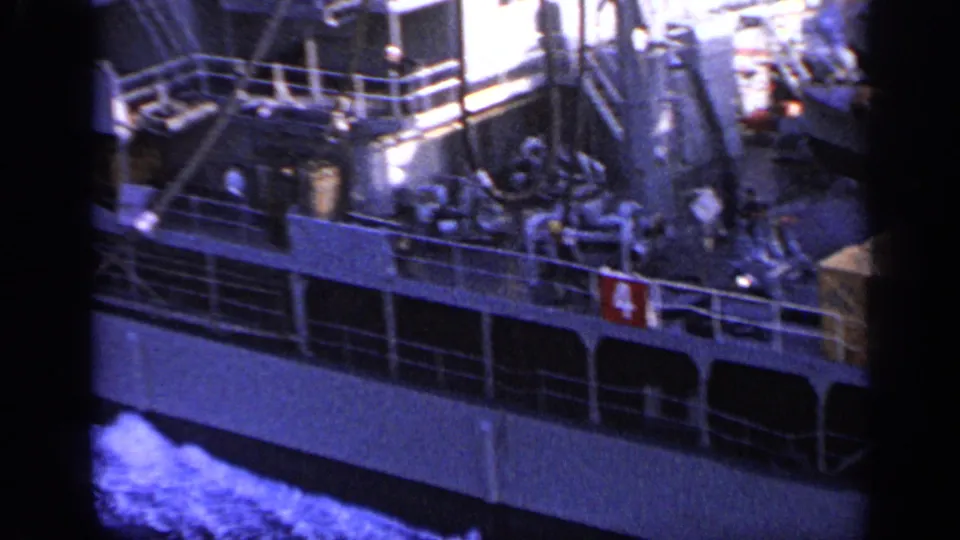 1962 home-movie frame showing crew figures on a cargo ship deck near Italy, with railings, ship structures, and equipment visible.