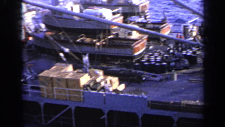 1962 8mm view of a cargo ship deck near Italy with stacked wooden boxes, dark barrels, railings, rigging, and a small deck craft.