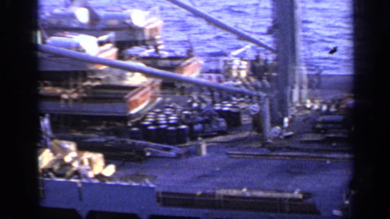Cargo ship deck with machinery, barrels, railings, and water beyond.
