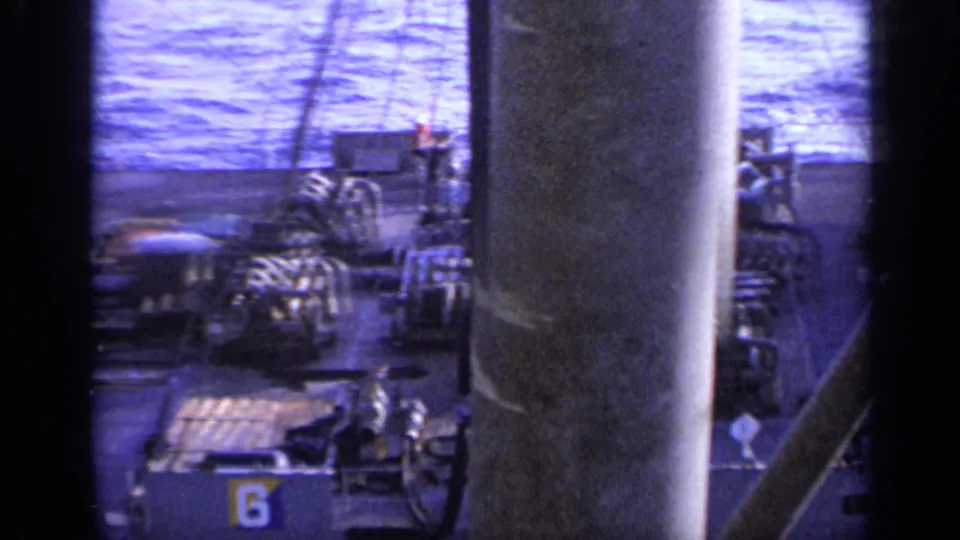 Blue-tinted 8mm frame showing shipboard equipment, pipes, a large column, water beyond, and a visible number marking on deck.