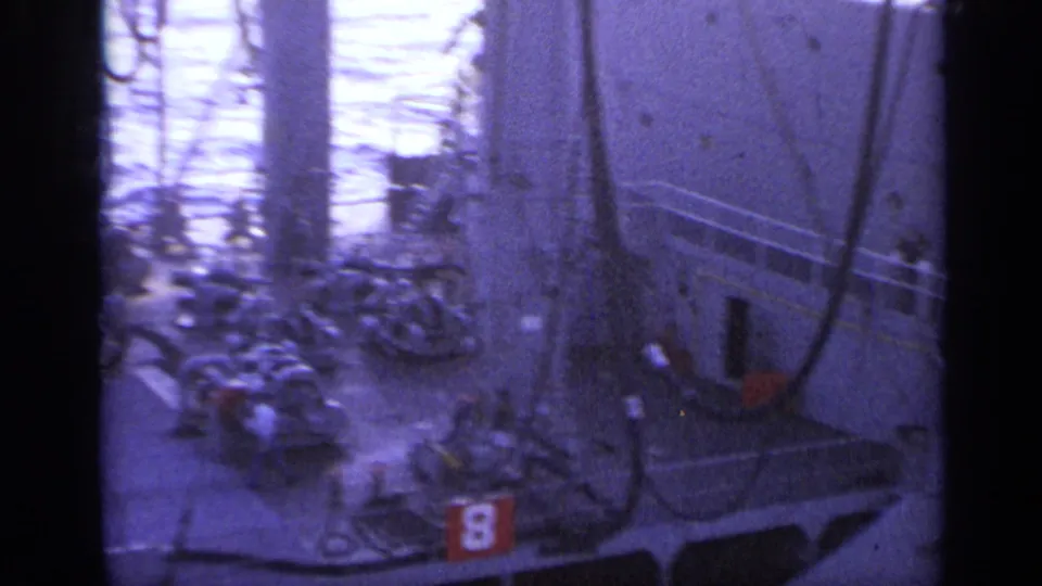 Blue 8mm view of crew activity around shipboard machinery, a large structural wall, railings, ropes, and a red numbered marking.