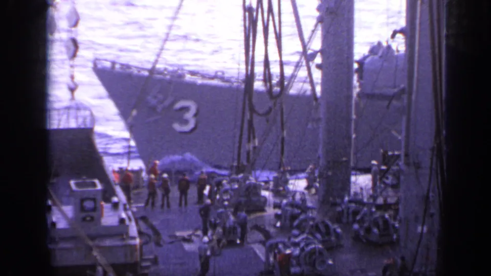 1962 Italy home-movie frame with a gray ship bow marked 3, people on deck, ropes, small craft, and bright water behind.