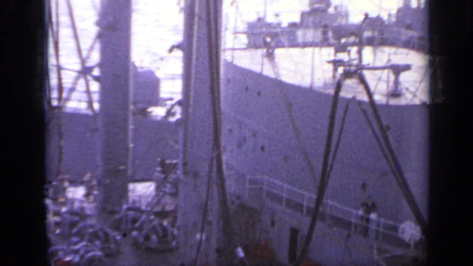 Soft 8mm frame showing the side of a gray naval vessel, foreground rigging, deck equipment, and small figures near the rail.