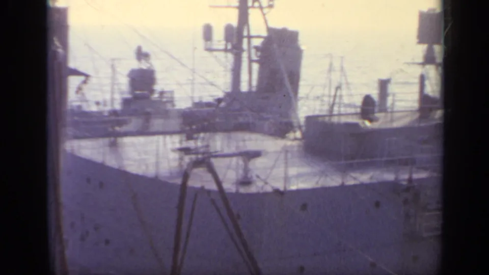1962 8mm frame with a gray ship deck in the foreground, a second vessel bow beyond it, rigging, railings, and bright water.