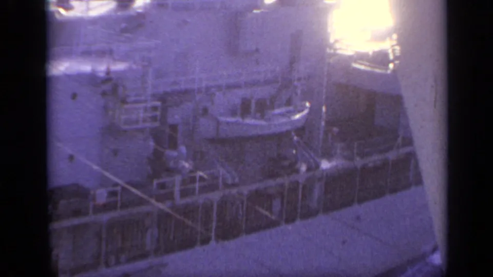 A close 8mm view of a naval ship at sea, showing railings, deck fittings, and bright water in the background.