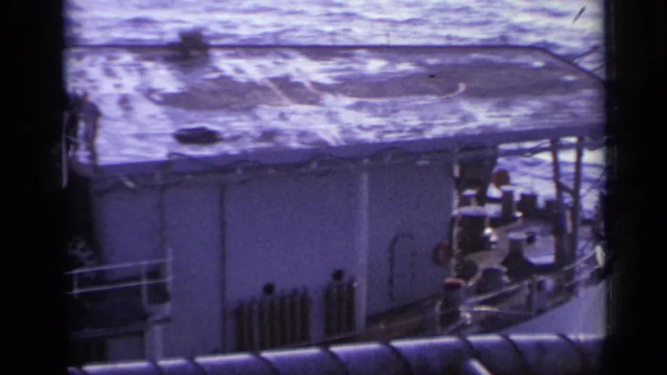 1962 home-movie frame showing the side of a naval vessel, ocean wake, railings, and small people near the deck edge.