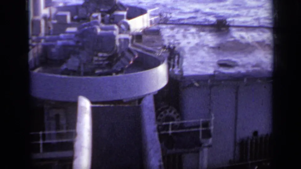 Aged 8mm frame from 1962 Italy showing a naval gun turret, deck equipment, railings, and ocean beyond.