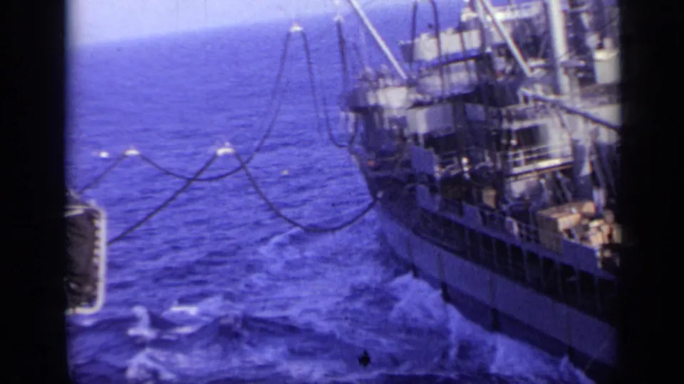 1962 Italy 8mm frame of a vessel's stern area, with deck structures, hanging lines or hoses, blue sea, and wake behind it.