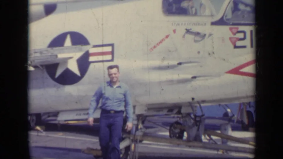 Man in a blue shirt stands on a ship deck in Italy in 1962 beside a U.S. Navy jet with star insignia.