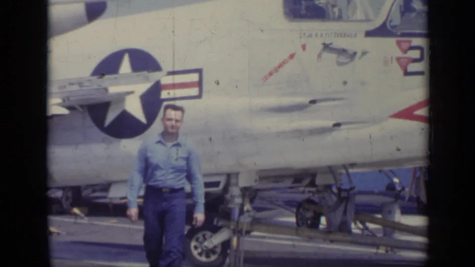 1962 Italy home-movie frame of a man facing the camera beside a Navy jet on a ship deck.