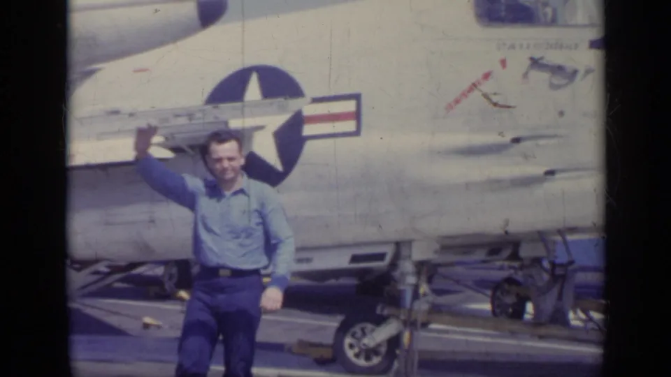Man waves toward the camera beside a U.S. Navy jet on a ship deck in Italy, 1962.