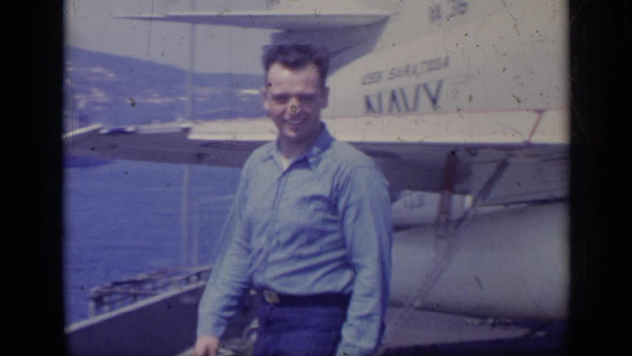 Smiling man stands near U.S. Navy-marked aircraft equipment with water behind him.