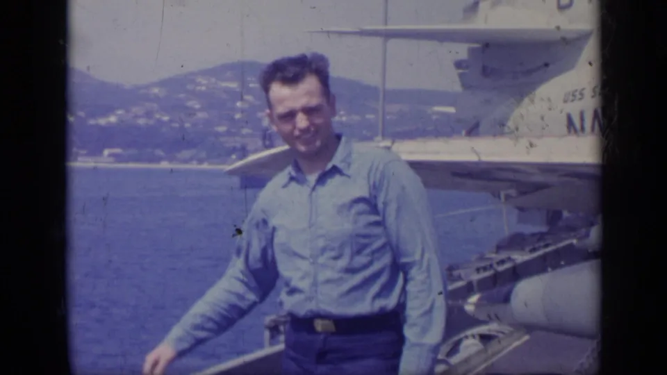 A man in a blue shirt poses on the USS Saratoga deck, with naval equipment, open water, and Italian coastline behind him.