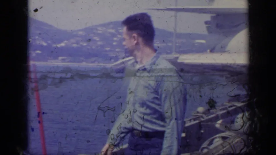 The man turns away from the camera in profile, standing near Navy aircraft hardware with the sea and distant shoreline visible.