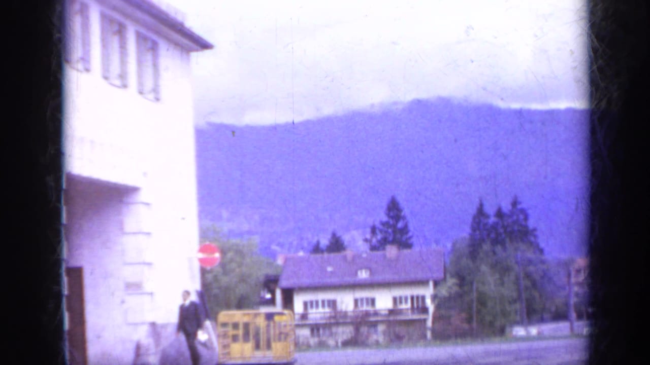 White building at left, distant mountains, yellow bus, red stop sign, and a suited pedestrian on a quiet street.