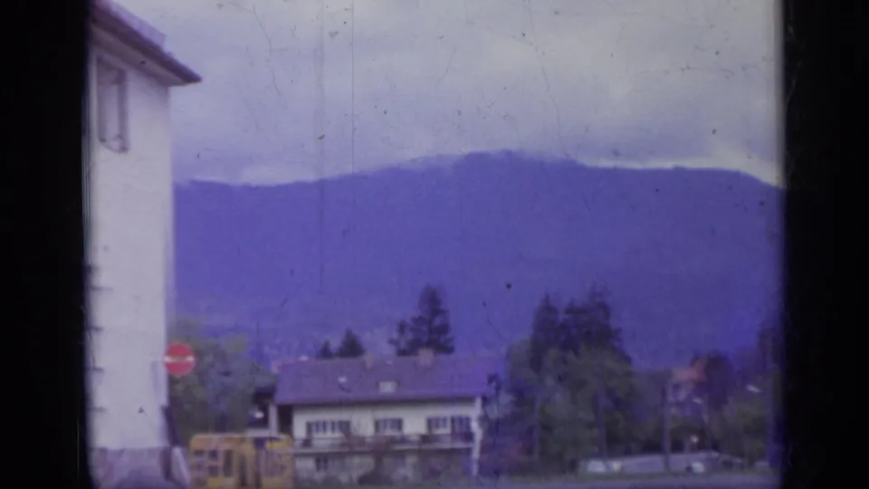 1962 Italy street frame with a white building at left, yellow bus near a stop sign, low houses, trees, and purple-tinted mountains under cloud.