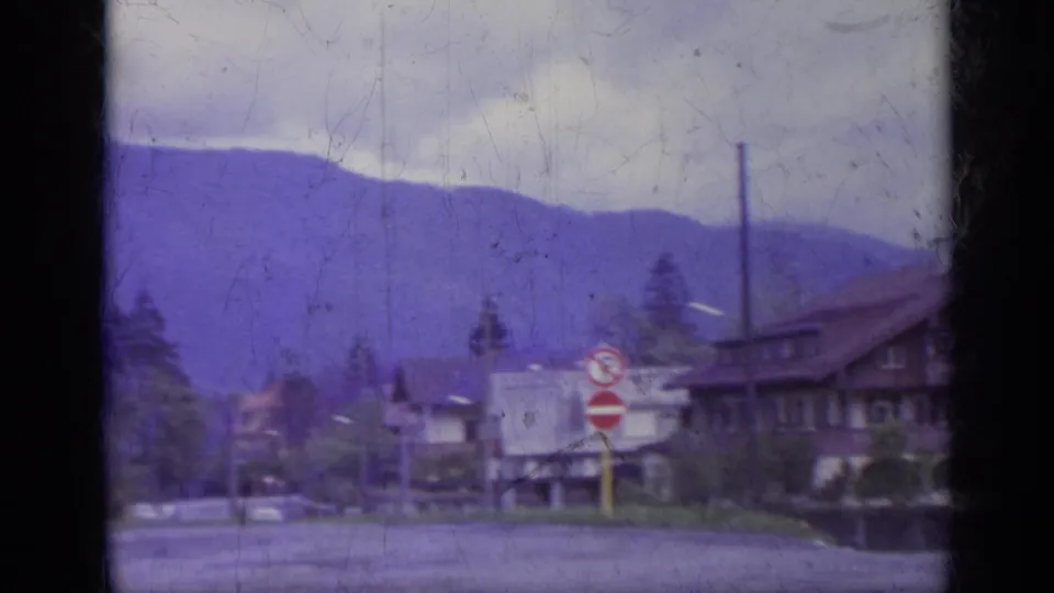 1962 Italy 8mm view across a road toward traffic signs, utility pole, low buildings, trees, and a dark-roofed residence below mountains.