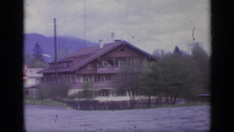1962 Italy frame showing a large chalet-style residence or lodge across the road, with trees, utility poles, mountains, and overcast sky.