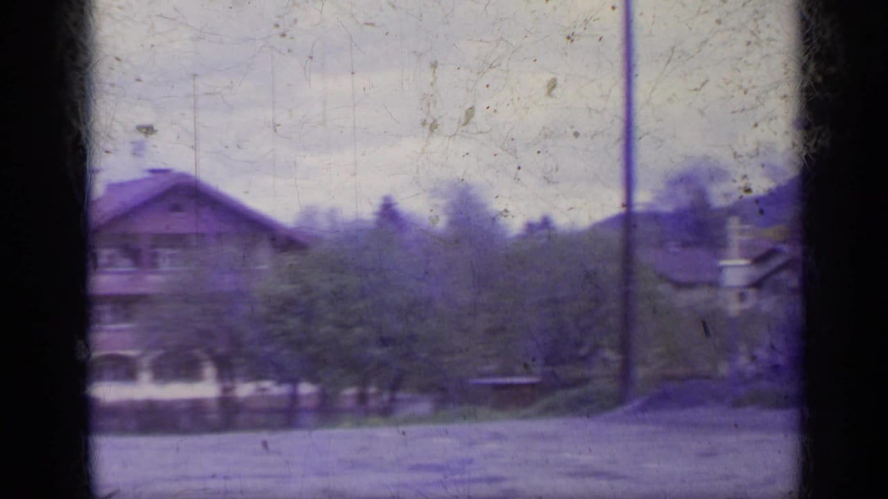 1962 Italy home-movie frame with a chalet-style building, trees, and open roadside space under a pale sky.