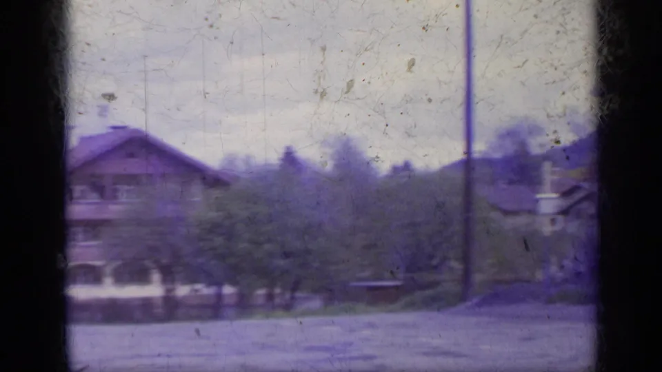 1962 Italy home-movie frame with a chalet-style building, trees, and open roadside space under a pale sky.