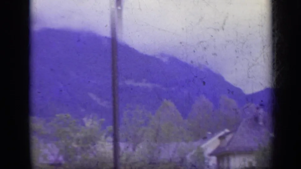 1962 Italy alpine frame with a mountain ridge, roadside trees, a utility pole, and part of a chalet-style roof.