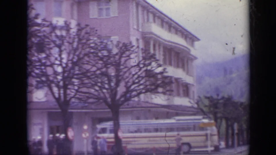 1962 Italian street scene with a multi-story building, pruned trees, a yellow coach bus, cars, and small distant pedestrians.