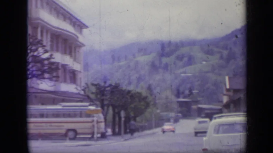 1962 Italian hillside road with a coach bus, classic cars, street trees, buildings, and green mountains beyond.