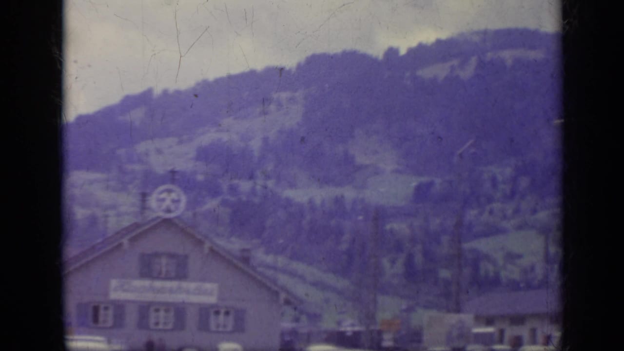 Chalet-style building below forested hills, partly blocked by a window frame.