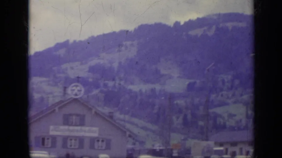 Italian alpine building and parked cars beneath wooded hills, seen through the divided frame of aged 8mm film.