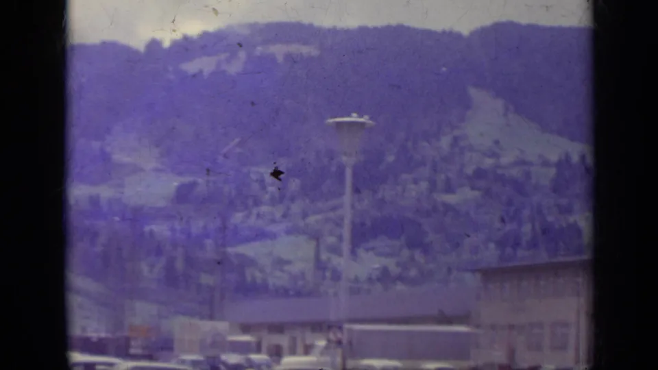 1962 Italian mountain town view with rooftops, a tall streetlamp, parked cars, and forested slopes in the background.