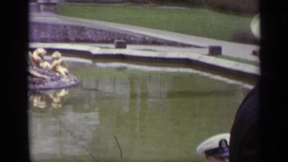1962 Italy 8mm frame looking across a fountain pool toward gilded sculpture, stone edging, lawn, and a partly visible military cap.