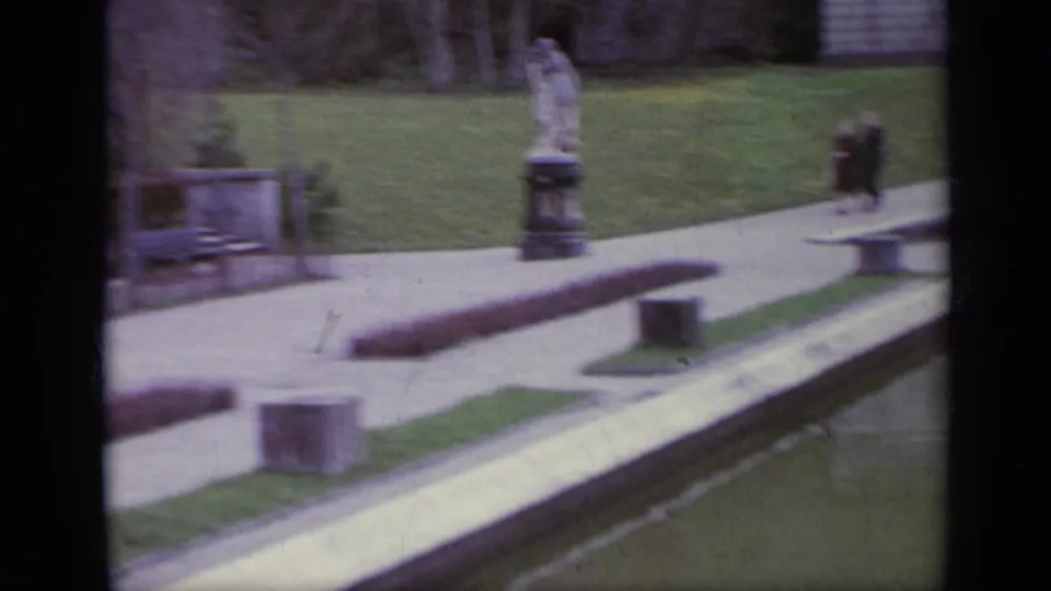 1962 Italy 8mm frame showing a garden walkway beside the pool, with statues, lawn, stone borders, and a distant person in dark clothing.