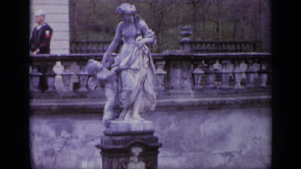 1962 Italy home-movie frame showing a classical statue, stone terrace, and a uniformed sailor near a historic building.