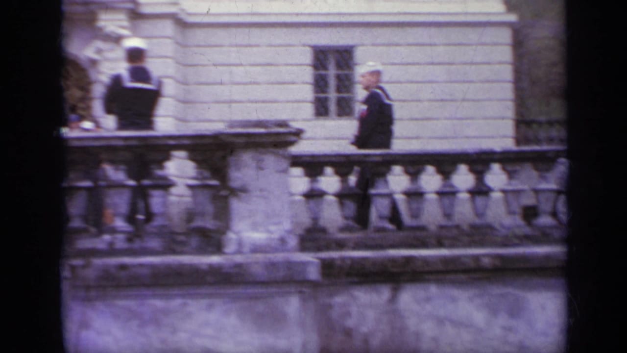 Two sailors stand near a stone railing in front of a pale historic building.