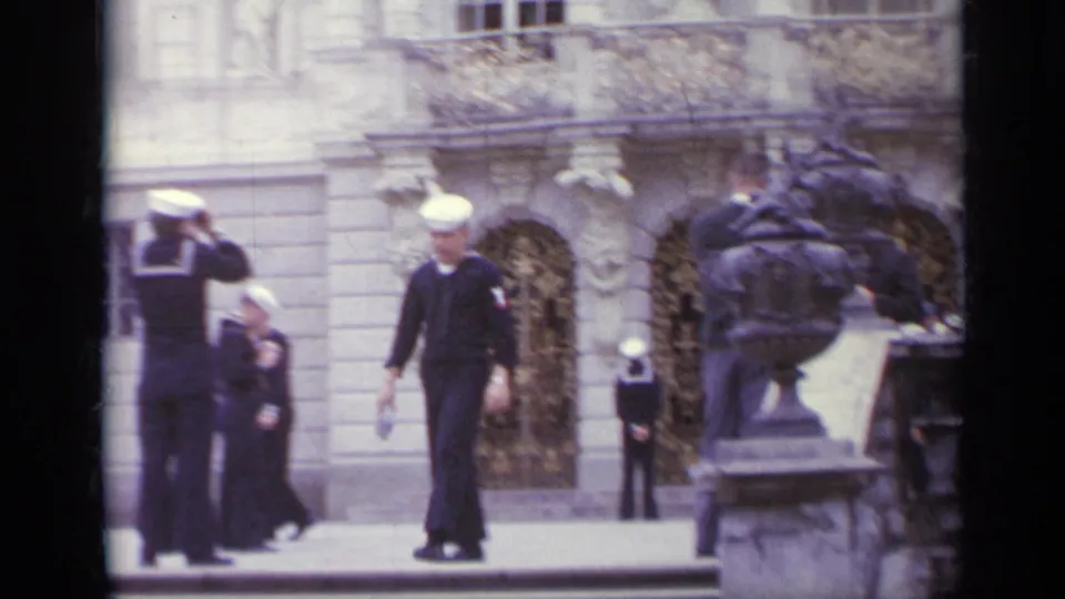 1962 Italy frame with uniformed sailors moving through a historic courtyard near a sculptural figure and decorated entrance.
