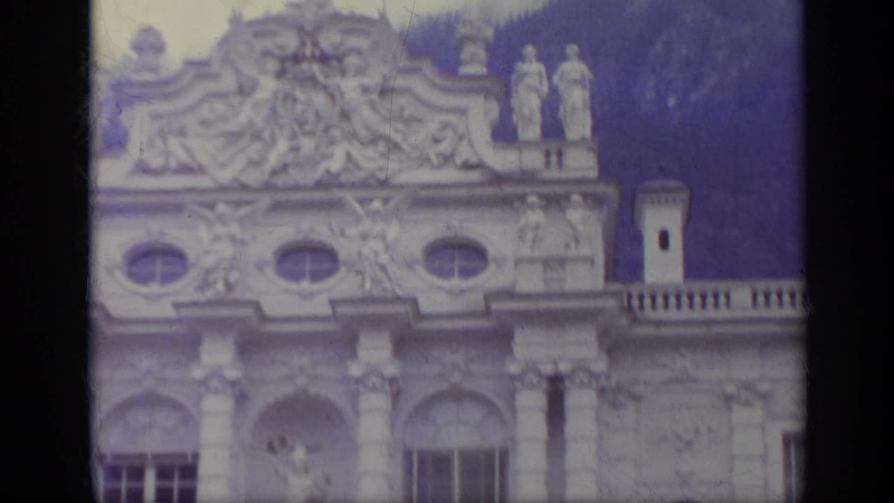 Ornate palace facade with sculpted figures and oval windows below a wooded hillside.