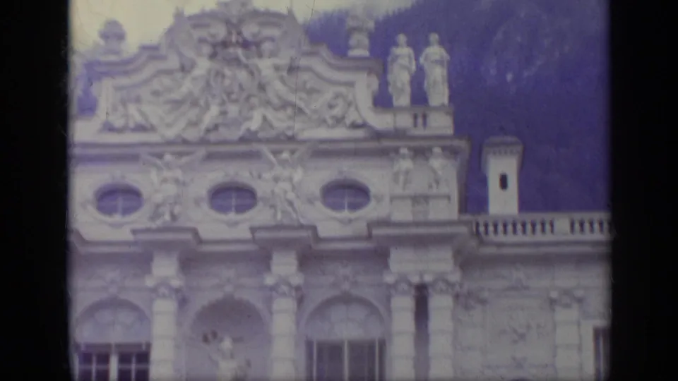 1962 Italy 8mm frame showing an ornate historic palace facade with statues, carved stonework, oval windows, and a dark wooded slope behind it.